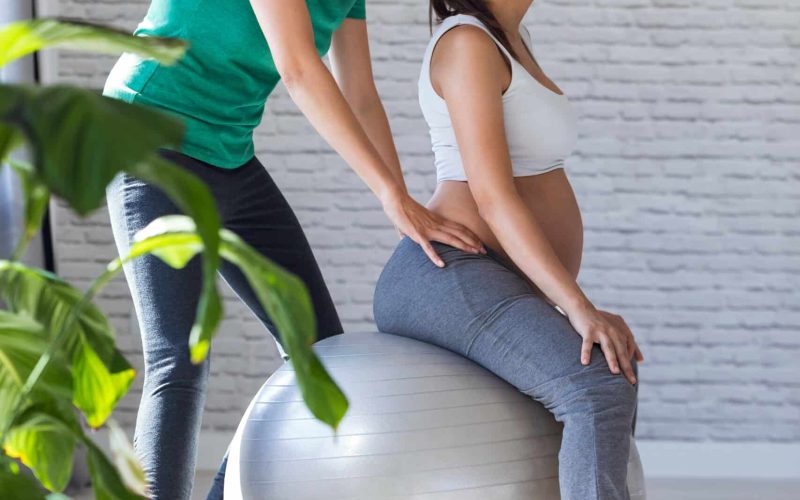 Shot of physiotherapist helping to beautiful pregnant woman for doing pilates exercises with ball preparing for childbirth.
