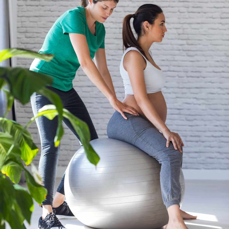 Shot of physiotherapist helping to beautiful pregnant woman for doing pilates exercises with ball preparing for childbirth.