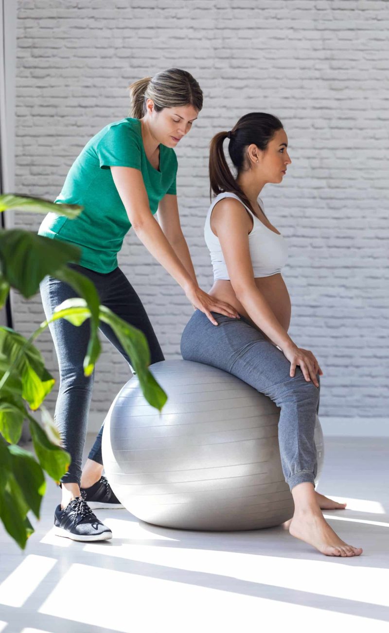 Shot of physiotherapist helping to beautiful pregnant woman for doing pilates exercises with ball preparing for childbirth.